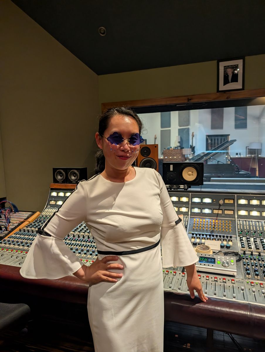 Michelle in a white dress at the mixing console, hand on hip, wearing the teardrop sunglasses.
