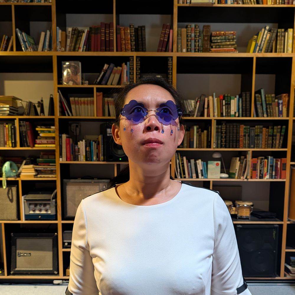 Michelle in teardrop sunglasses, not smiling, in front of the studio bookshelves.